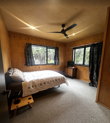 Dieses gemütliche Schlafzimmer verfügt über Holzwände, ein Doppelbett, zwei große Fenster mit gemusterten Vorhängen, einen Deckenventilator, einen kleinen Schreibtisch mit Monitor und einen Teppichboden in neutralen Farbtönen.
