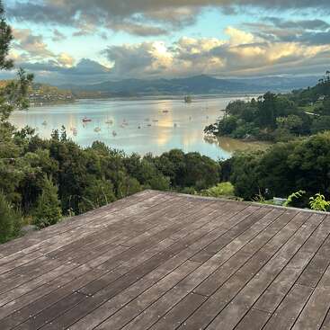 Una terraza de madera da a una tranquila bahía con veleros, rodeada de verdes colinas y árboles bajo un espectacular cielo nublado al atardecer, bañado por una suave luz solar.