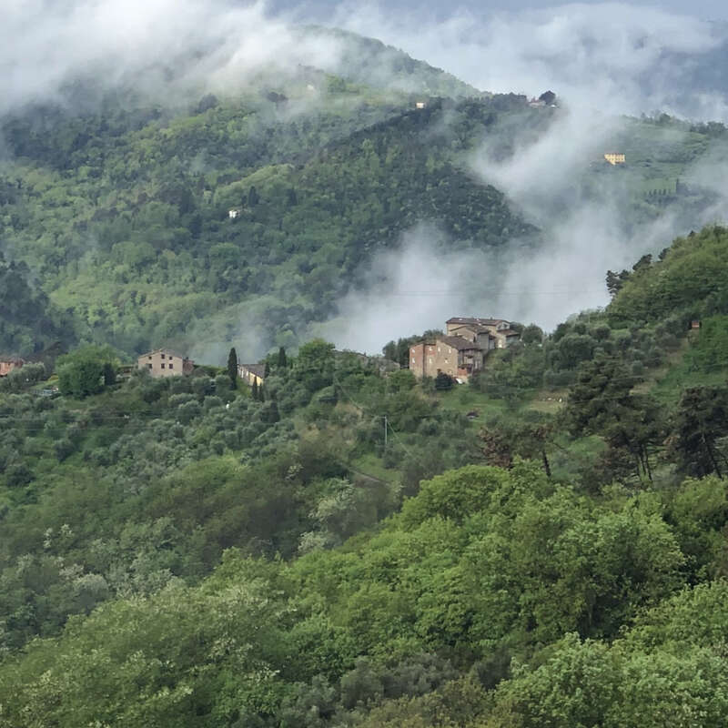 The image depicts a serene mountainous landscape with lush greenery, featuring a few buildings nestled among the trees, set against a cloudy sky.