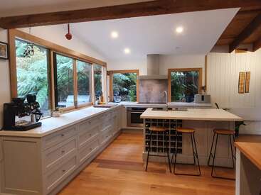 Esta cocina cuenta con amplios ventanales con vistas al bosque, armarios blancos, suelos de madera, una isla con taburetes, una cafetera, electrodomésticos modernos y una decoración elegante y minimalista.