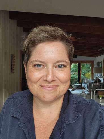 A woman with short hair smiles warmly at the camera indoors. She wears a dark collared shirt, with natural light and a cozy home in the background.