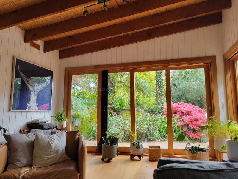 Un acogedor salón con vigas de madera a la vista, grandes puertas acristaladas, plantas de interior, cómodos asientos y unas preciosas vistas al jardín, con un follaje de vivos tonos verdes y rosas.