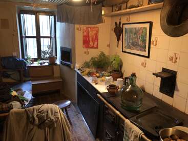 In dieser gemütlichen, rustikalen Küche sorgen ein Holzofen, Pflanzen auf der Fensterbank, Vintage-Dekor, verstreute Kleidung und verschiedene Küchenutensilien für eine warme, lebendige Atmosphäre.