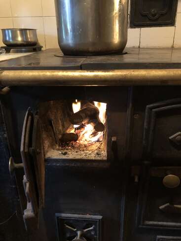 Die Tür eines Holzofens, in dem ein Feuer brennt, ist geöffnet. Darauf stehen große Metalltöpfe, die auf traditionelles Kochen oder Heizen hindeuten.