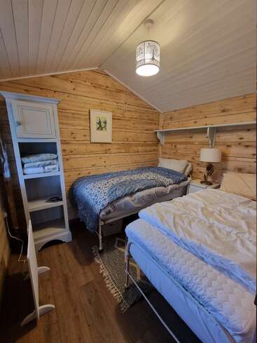 A cozy rustic bedroom with wooden plank walls, two single beds, a white cabinet, a small lamp, wall shelf, and a picture hanging above the bed.