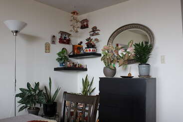 Um cantinho aconchegante com plantas domésticas, uma cômoda preta, prateleiras flutuantes com estatuetas, um espelho oval, decoração suspensa, um abajur de pé e uma cadeira de madeira.