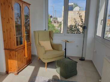 Um cantinho aconchegante pra leitura com uma poltrona verde, pufe combinando, armário de madeira, mesinha lateral e luminária de pé perto de janelas grandes e ensolaradas, criando um clima tranquilo.