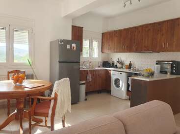 Una acogedora cocina con armarios de madera, una mesa redonda con sillas, una nevera gris, lavadora, microondas, frutero y ventanas soleadas que proporcionan luz natural.