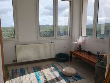Una acogedora habitación en esquina con grandes ventanas con vistas a verdes colinas, radiador, alfombra a rayas, cojín en el suelo, cama pequeña, banco de madera y jarrón decorativo. Ambiente tranquilo.