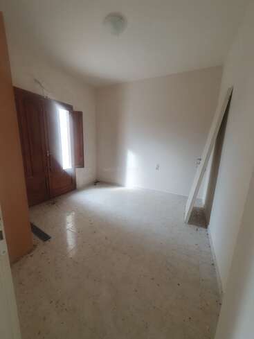 Es una habitación pequeña y vacía, con paredes blancas, suelo de baldosas sucias, contraventanas de madera, poca luz natural y una puerta apoyada contra la pared.
