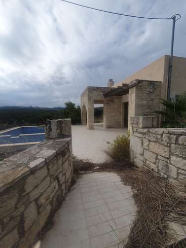Un camino de piedra conduce a una villa rústica con paredes y arcos de piedra, con vistas a las montañas. Las plantas crecidas y el cielo nublado crean una atmósfera mediterránea abandonada y serena. Se ve una piscina azul.