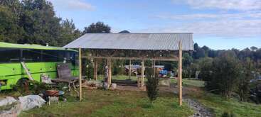 A simple open wooden structure with a metal roof sits on grassy land. A bright green bus, trees, tools, and various objects surround the rustic scene.