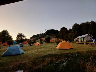 The image shows a peaceful campsite at dusk, featuring multiple colorful tents on grassy land, a bicycle, surrounding trees, and a small building with laundry hanging outside.
