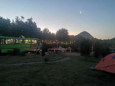 At dusk, a campsite with a green bus, string lights, picnic tables, and tents creates a cozy outdoor gathering, framed by trees, mountains, and a crescent moon.