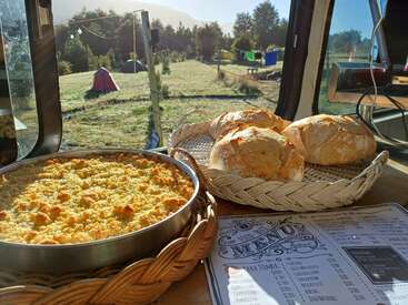 Fresh homemade bread and a crumbly casserole sit on a wooden table by a window, overlooking a sunny campsite with tents and lush green scenery outside.