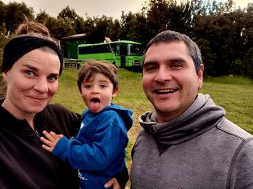 A family of three smiles outdoors. The child playfully sticks out his tongue. Behind them is a bright green bus, surrounded by grass and trees.