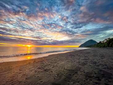 Golden sunlight illuminates a tranquil beach with dark sand, gentle waves, vibrant clouds, and a distant mountain. Footprints suggest recent visitors, enhancing the scene’s peaceful beauty.