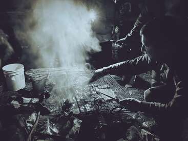 A group of people gather around a smoky outdoor grill at night, cooking meat over open flames. Shadows and light create a dramatic, atmospheric camping scene.