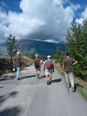 Um grupo de pessoas caminha por uma estrada rural cercada por árvores e montanhas sob um céu parcialmente nublado, aproveitando a natureza e a companhia uns dos outros ao ar livre.