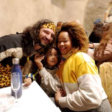 Una familia sonriente se sienta junta en un acogedor restaurante, vestida con coloridos trajes. El niño mira hacia arriba con cariño. Ambiente cálido, trajes festivos y expresiones alegres por todas partes.