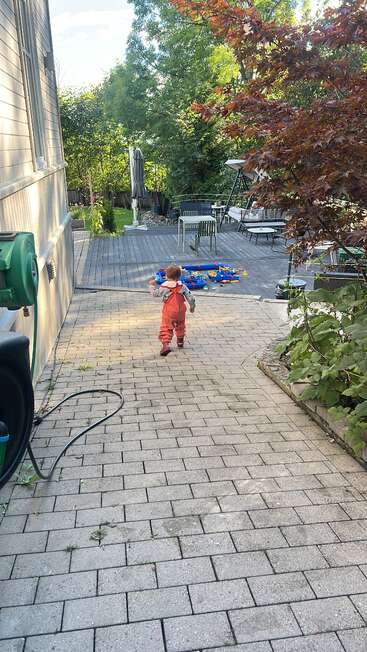 Un niño con mono rojo camina por un sendero de piedra hacia una terraza con juguetes, muebles de jardín y exuberante vegetación en un día soleado.