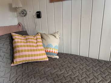 Una cama acogedora con una manta acolchada gris, dos cojines decorativos -uno a cuadros y otro geométrico- contra una pared de paneles blancos. Se ven una lámpara de pared y un cargador.