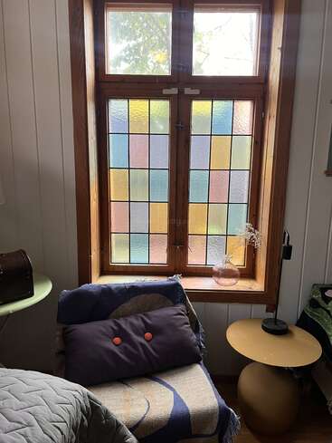 This cozy corner features a colorful stained glass window, a patterned armchair with a pillow, a round side table, and a decorative vase with dried flowers.