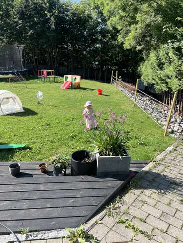Un patio trasero soleado con un niño con un gorro rosa, juegos infantiles, un tobogán, macetas en una terraza de madera, césped verde y árboles a su alrededor. Una escena tranquila.