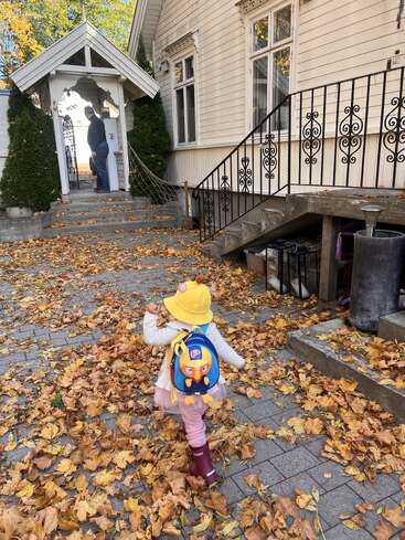 Un niño pequeño, con un sombrero amarillo y una mochila, camina entre las hojas caídas del otoño hacia una casa blanca, donde un adulto le espera en la entrada.