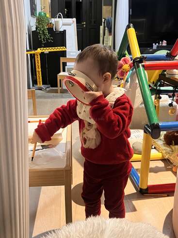 Un niño pequeño vestido de rojo se tapa la cara en tono juguetón con una pandereta, de pie junto a una mesita de madera en una habitación acogedora y colorida.
