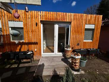 Cette image montre une petite cabane en bois avec des lambris verticaux, des portes coulissantes en verre, des jardinières, deux fenêtres, des décorations extérieures et un porche en bois donnant sur un jardin.