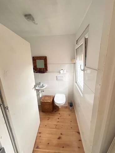 Cette photo montre une petite salle de bains toute simple, avec un sol en bois, des toilettes suspendues, un petit lavabo, un panier en osier, un miroir, une brosse de toilettes et une fenêtre.