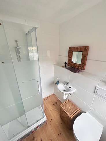 Cette salle de bains est équipée d'une douche vitrée, d'un petit lavabo, de toilettes blanches, d'un parquet, d'un panier en osier et d'un miroir décoratif au-dessus du lavabo, créant ainsi une ambiance chaleureuse et minimaliste.