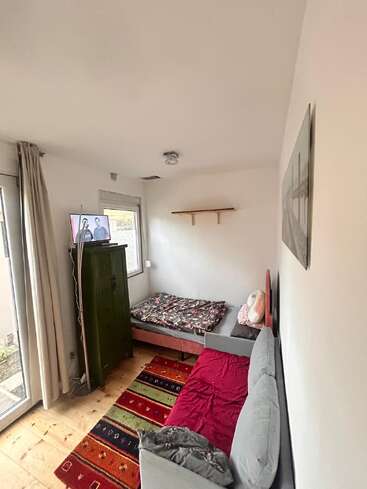 Une petite pièce chaleureuse avec un lit, un canapé gris, un tapis coloré, une armoire verte, une télévision fixée au mur, une fenêtre, des étagères et une décoration minimaliste. Sol en bois clair.