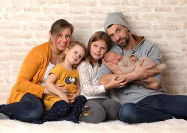 L'image représente une famille de cinq personnes assise sur un tapis blanc devant un mur de briques blanches, les parents tenant leur nouveau-né et leur petite fille.