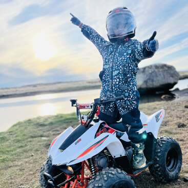 The image depicts a young child standing on an ATV, pointing to the left with their right hand and giving a thumbs-up with their left hand.