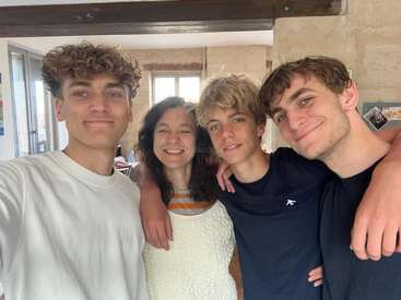 Four people, three young men and one woman, stand closely together indoors, smiling happily. Their arms are around each other, showing warmth and family affection.