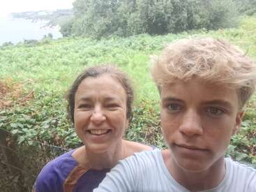 A woman and a boy take a selfie outdoors, smiling in front of lush greenery with trees and a distant seaside cliff visible in the background.