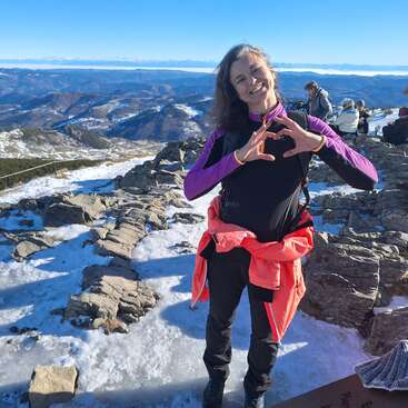 A woman stands on a snowy mountain, smiling and making a heart shape with her hands. The sun shines brightly, and stunning mountains stretch behind her.