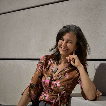 A woman with dark hair, wearing a brown floral blouse, sits and smiles against a light-colored concrete wall, enjoying sunlight with a relaxed, cheerful demeanor.