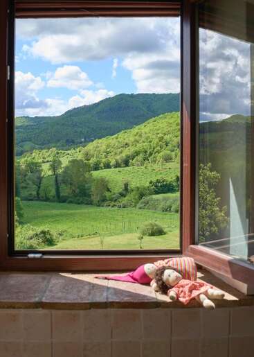 A wide open window overlooks lush green hills under a partly cloudy sky. Two handmade cloth dolls rest on the sunlit windowsill, creating a serene scene.