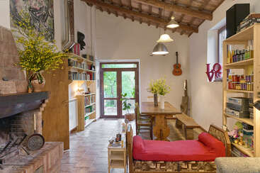 A cozy rustic living room features wooden ceilings, a brick fireplace, bookshelves, a wooden dining table, red sofa, guitar on the wall, and vibrant yellow flowers.