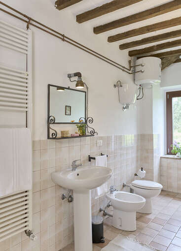 This rustic bathroom features exposed wooden ceiling beams, cream tiles, a pedestal sink, bidet, toilet, mirror, towel rack, water heater, shelf, and window.