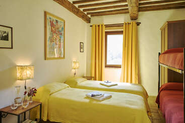 This cozy bedroom features two single beds with yellow bedding, a bunk bed, rustic wooden beams, yellow curtains, warm lighting, artwork, and a window view.