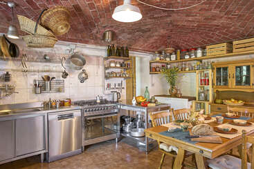 This warm rustic kitchen features a brick vaulted ceiling, stainless steel appliances, open shelves with jars, wooden dining table set for a meal, and hanging baskets.