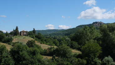 This image shows a peaceful countryside landscape with a house on a hill, surrounded by lush green trees, rolling fields, distant hills, and a bright blue sky.