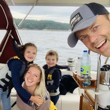 The image shows a family of four on a boat, with the father wearing a baseball cap and smiling at the camera, surrounded by his loved ones.