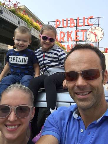 L'image montre une famille de quatre personnes posant pour un selfie devant un panneau indiquant "Marché public", avec une horloge et des fleurs en arrière-plan.