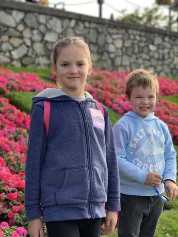 L'image montre deux enfants debout devant un mur de pierre et un jardin de fleurs roses, l'enfant de gauche portant un sweat à capuche bleu et l'autre un sweat à capuche bleu clair.