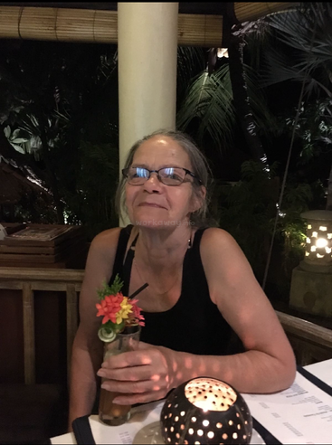Uma mulher de óculos sorri, segurando uma bebida decorada em um restaurante à luz de velas. O clima noturno com plantas tropicais e iluminação aconchegante cria uma atmosfera relaxante e agradável.